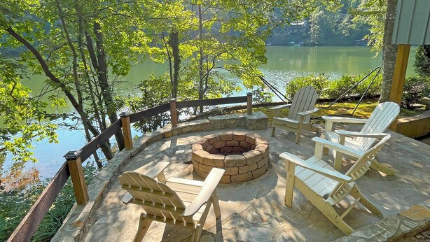 A deck with a stone fire pit and Adirondack chairs overlooking a lake.