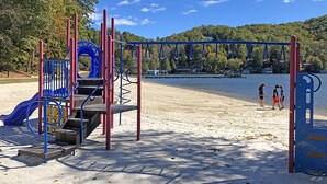 A playground with a slide and climbing structures, located near a beach with a clear view of the water and hills in the background.