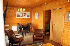 A wooden cabin interior with a dining area, two leather chairs, a chandelier, and a door leading to another room.