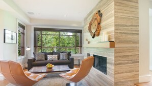 Una sala de estar moderna con un sofá, dos sillones de cuero, una mesa de centro con fruta, una chimenea y ventanas amplias con vistas a zonas verdes.