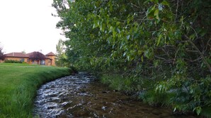 Un ruisseau traverse une zone herbeuse, bordée d’une végétation dense d’un côté, et on aperçoit un bâtiment au loin.
