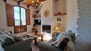 Una sala de estar acogedora con dos sofás, un televisor, una chimenea y un amplio ventanal con contraventanas de madera.
