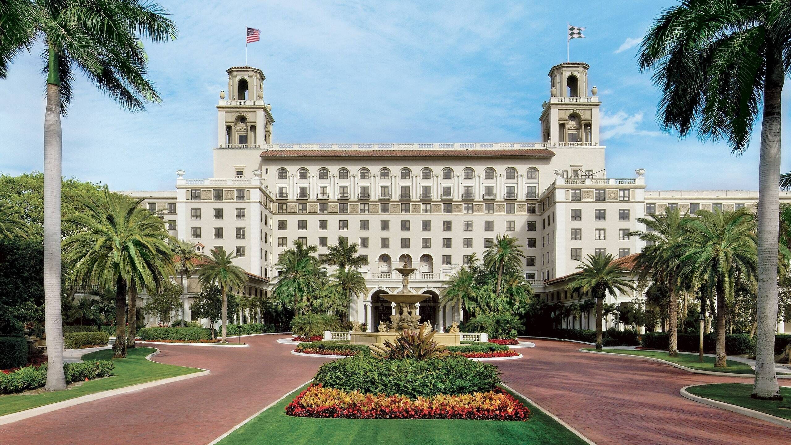 A large, historic building with two towers, a central fountain, and a well-maintained garden with palm trees.