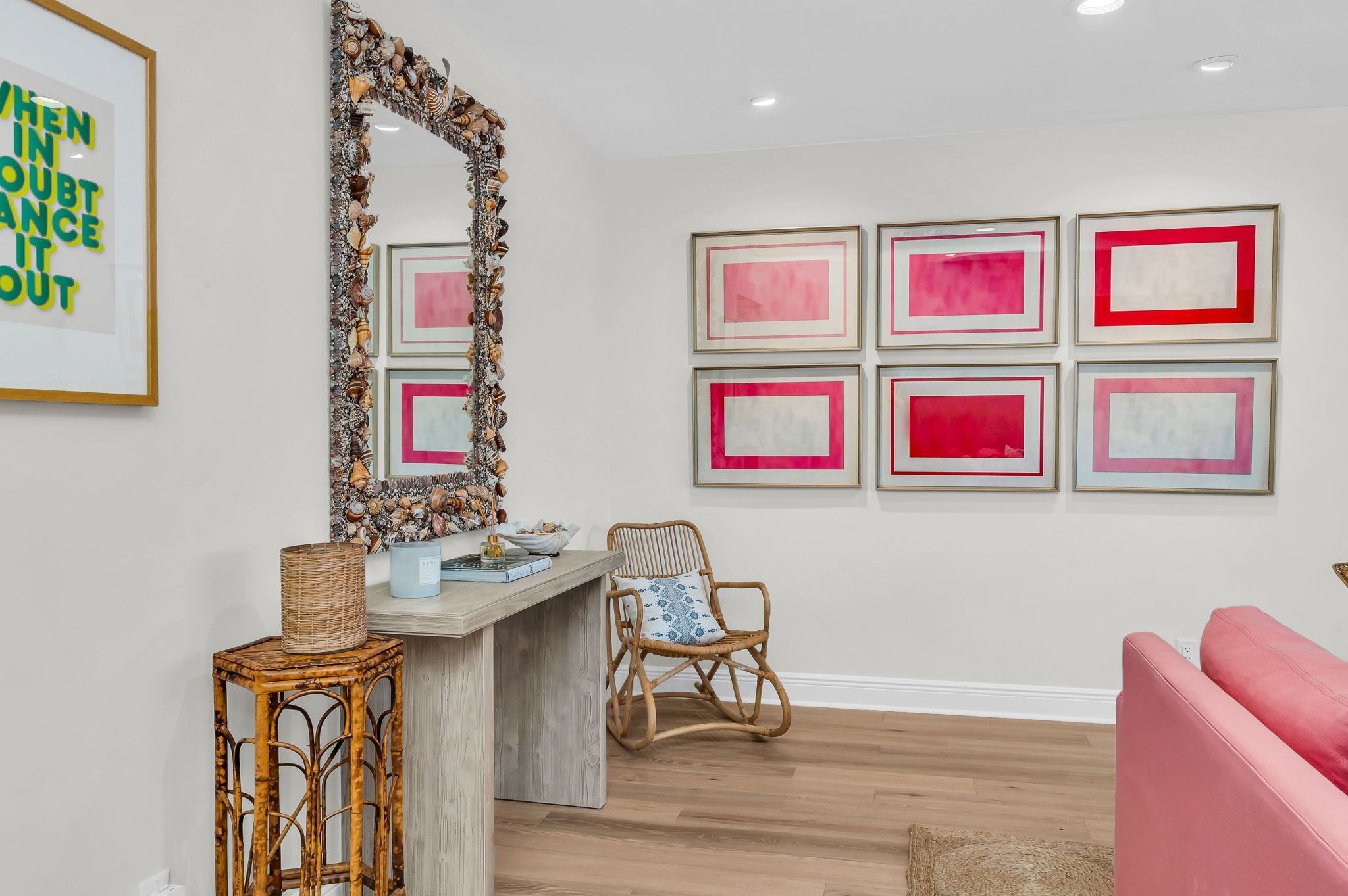 A room with a wooden desk, wicker chair, and a pink sofa. There are framed artworks on the wall and a decorative mirror with a seashell border.
