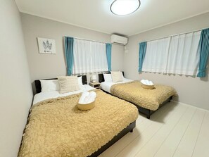 Une chambre d’hôtel avec deux lits, chacun recouvert d’une courtepointe beige à texture et agrémenté d’un panier de serviettes blanches. On y trouve un luminaire au plafond, un appareil de climatisation et une œuvre d’art encadrée au mur.