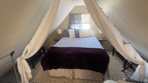 Ein Bett mit Baldachin, ein Nachttisch, eine Lampe und ein gemustertes Teppich in einem Raum mit schrägen Decken.