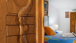 Une porte en bois au design original, un lit recouvert d’une courtepointe à motifs floraux colorés, une table de chevet blanche avec une lampe, et une porte en bois à l’arrière-plan.