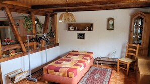 A cozy bedroom with a wooden ceiling, a bed with a patterned bedspread, a wooden chair, a small table, and a wall-mounted shelf with decorative items.