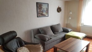 A living room with a grey sofa, a black armchair, a wooden coffee table, a green blanket, a painting on the wall, and a floor lamp.