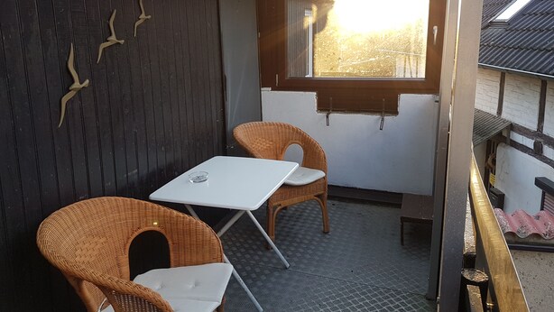 A small outdoor seating area with wicker chairs and a table, a window with a view of trees, and a dark wooden wall with decorative elements.