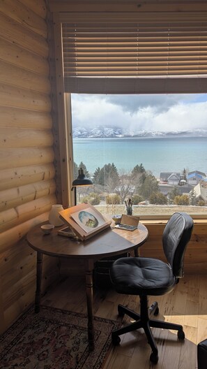 Una camera accogliente con arredamento in legno, una scrivania con una sedia, un quadro incorniciato e vista su un lago e montagne lontane.
