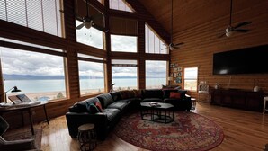 Un salon spacieux, doté d’un grand canapé d’angle, d’une table basse ronde et offrant une vue sur un lac grâce à de grandes fenêtres.