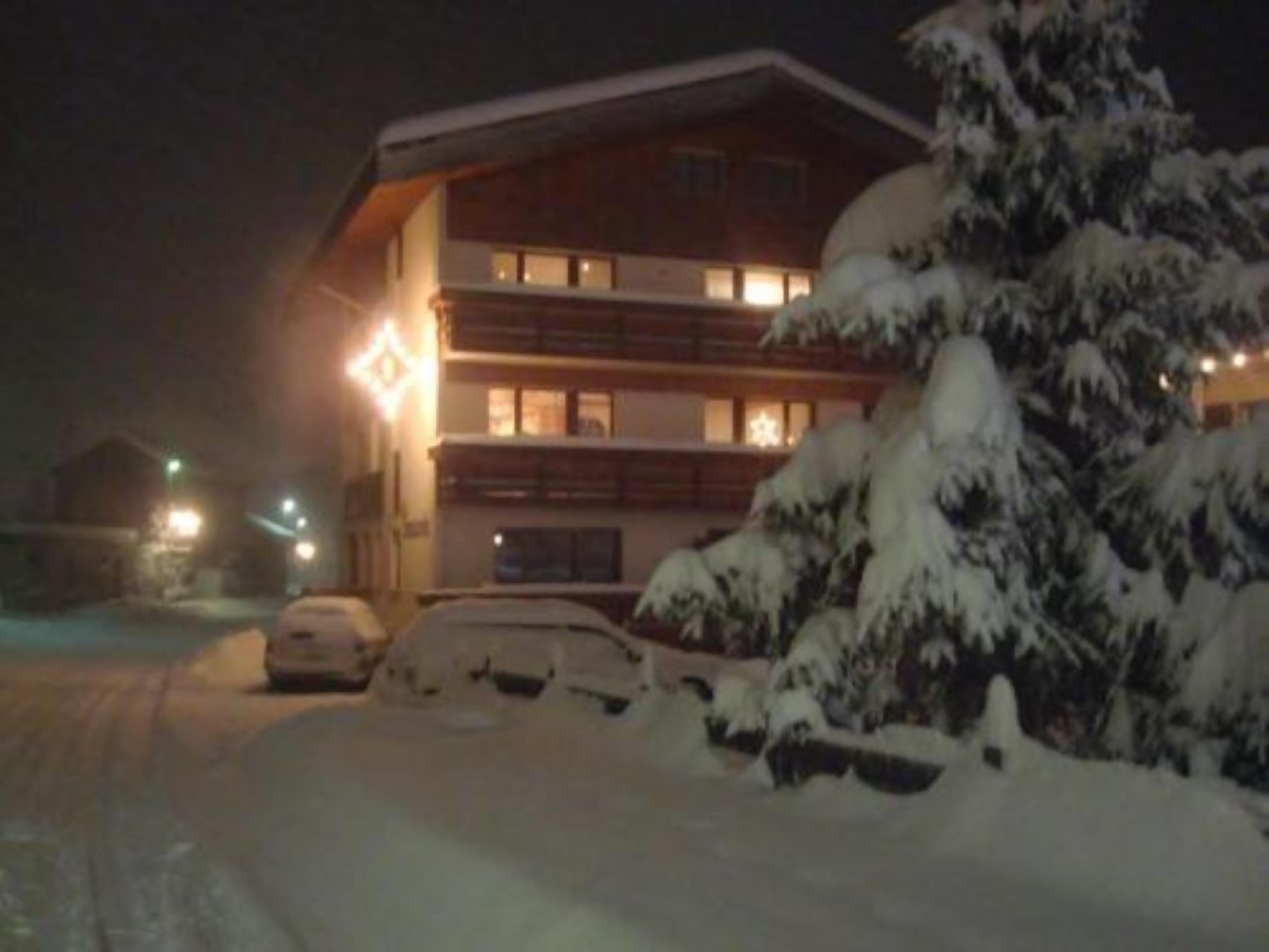 Um edifício de vários andares com paisagem coberta de neve, um carro estacionado e uma escadaria visível que leva à entrada.
