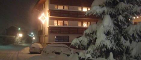 Um edifício de vários andares com paisagem coberta de neve, um carro estacionado e uma escadaria visível que leva à entrada.
