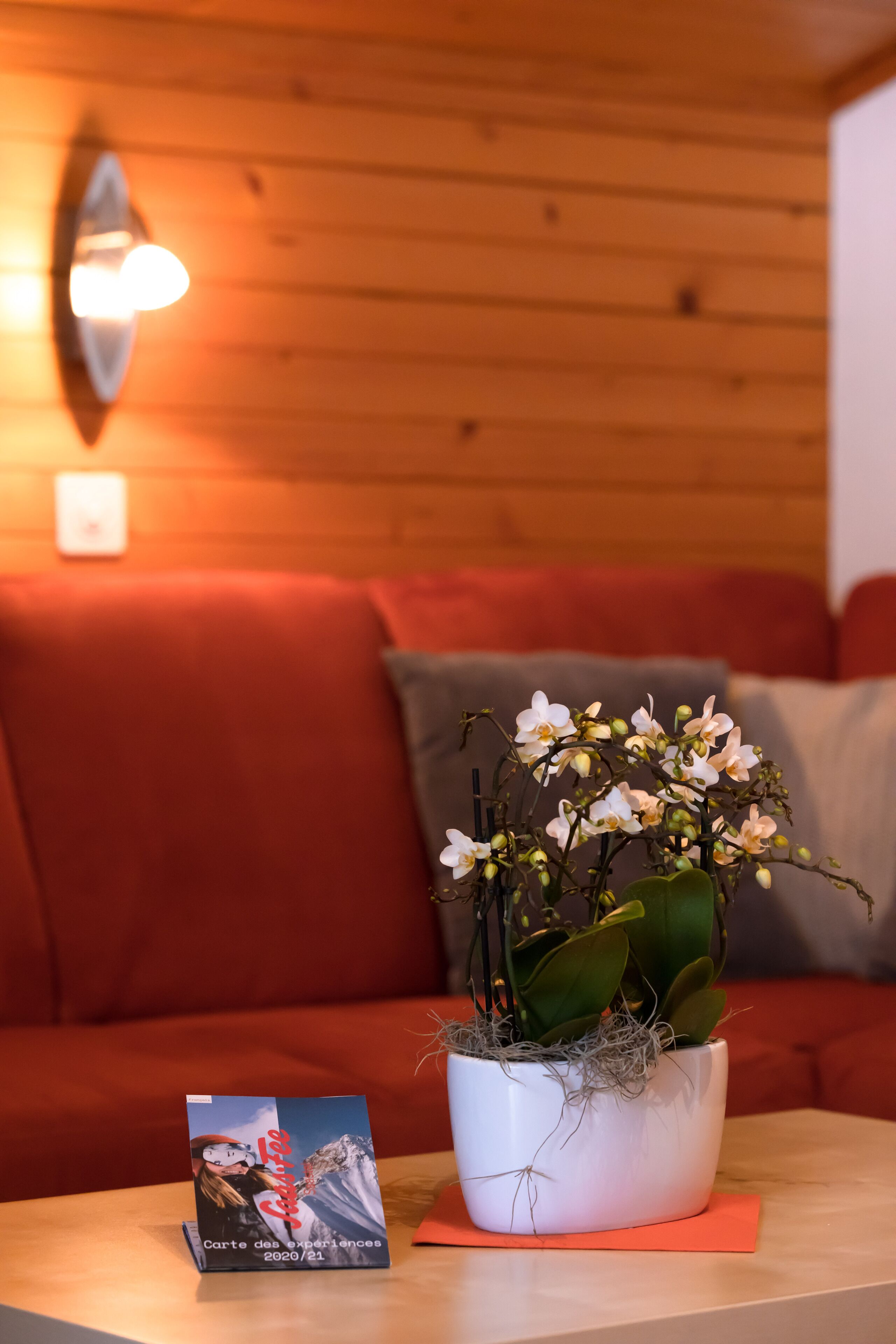Pared de madera con una lámpara, un sofá rojo, una orquídea blanca en maceta y un folleto sobre una mesa.
