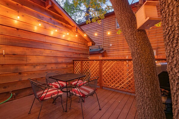 Eine Terrasse im Freien mit Holzwänden, Lichterketten, einem Metalltisch mit Stühlen und einem Holzzaun.