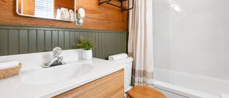 Un baño con tocador de madera, un lavamanos blanco, un inodoro con asiento de madera, cortina de ducha y un espejo.