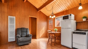 A room with a wooden floor, a gray sofa, a dining table with chairs, a kitchen area with a stove, a microwave, and a refrigerator.