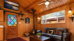 A cozy cabin interior with a fireplace, leather sofa, wooden floors, and a large window.