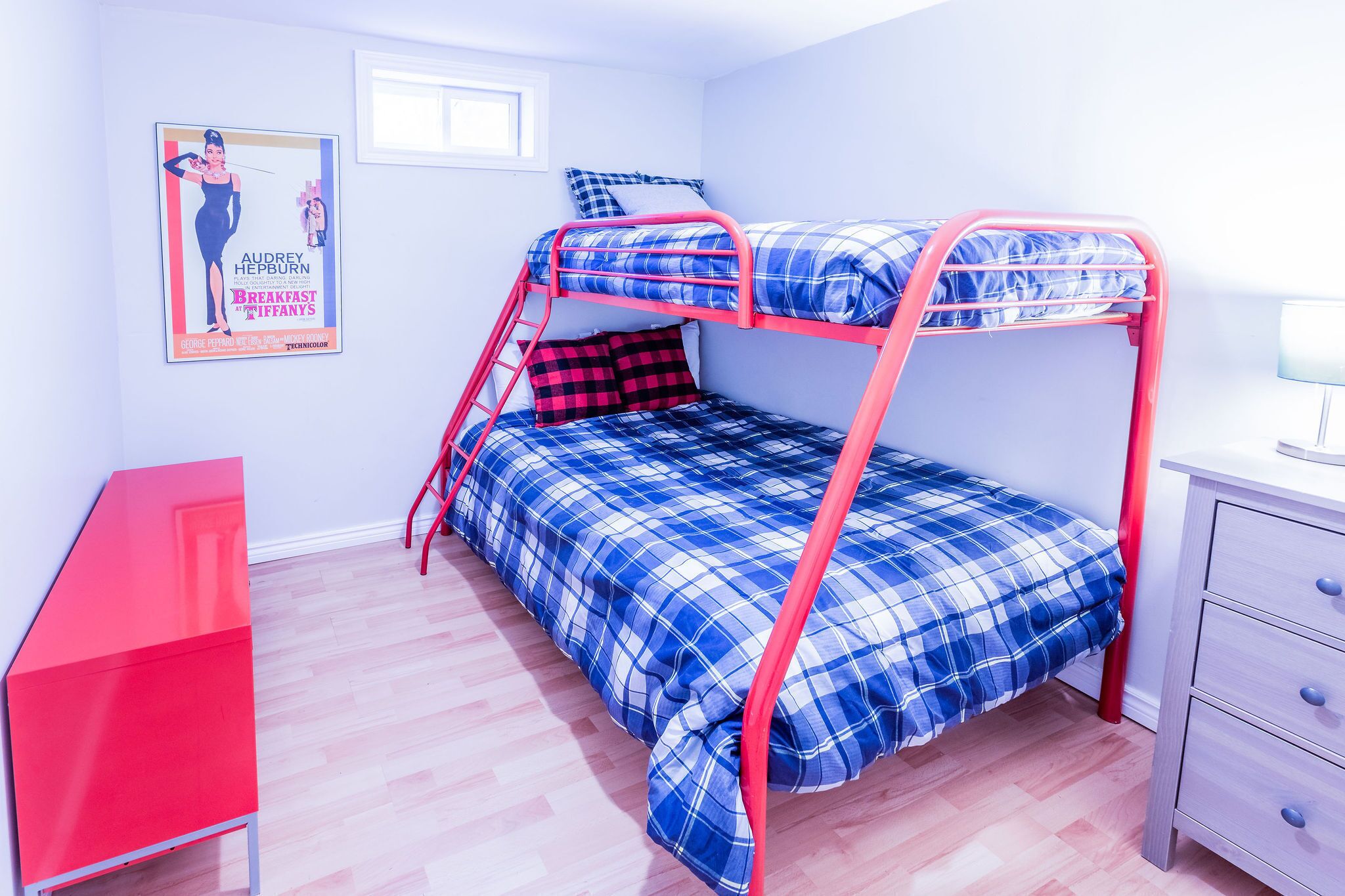 A bunk bed with a blue and red checkered bedspread, a red desk, and a small dresser.