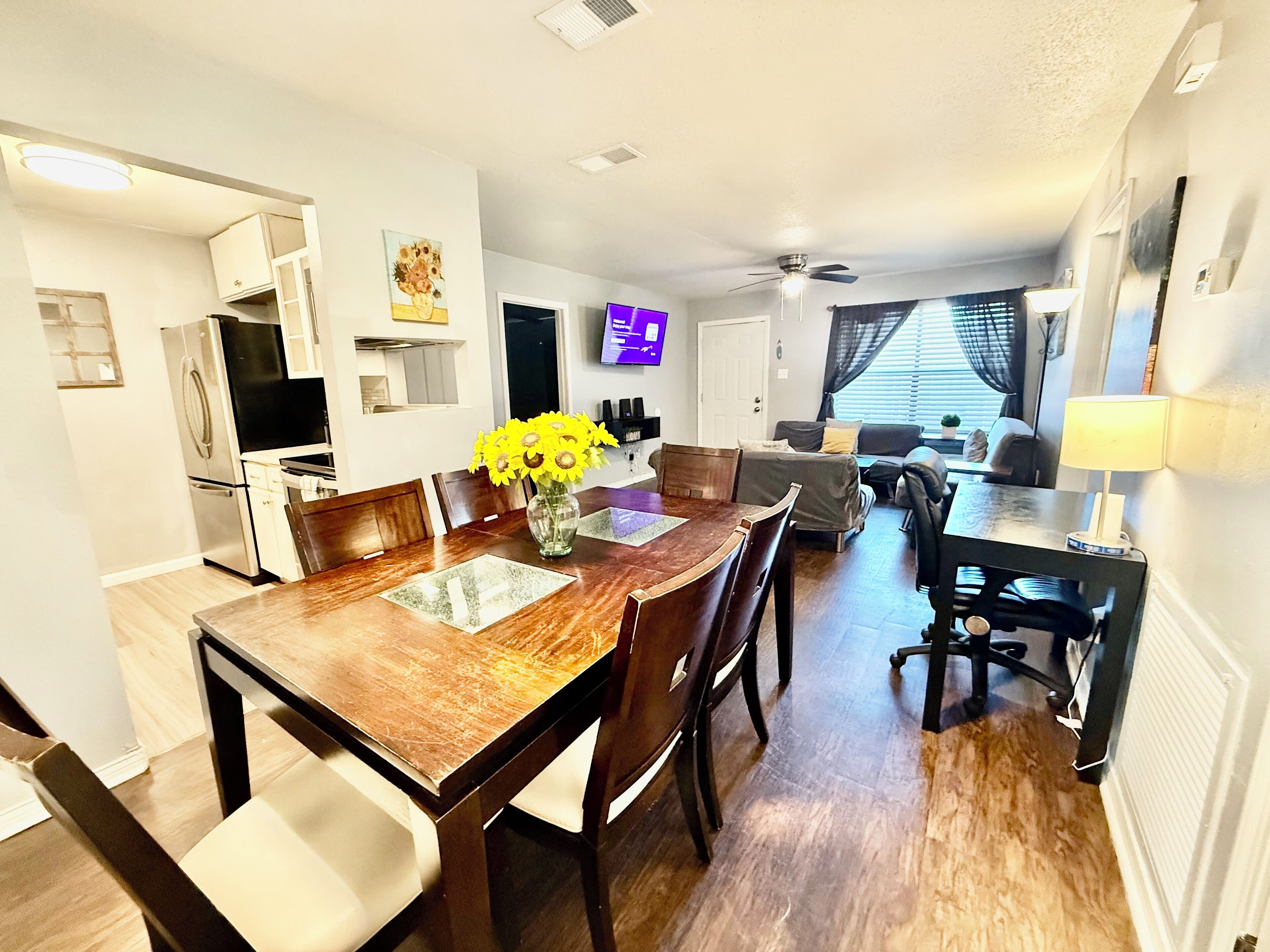 A modern kitchen with a dining area, a living room with a sofa and a TV, and a ceiling fan.
