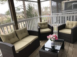 Een overdekt terras met rieten meubels en kussens, een glazen bijzettafeltje en een vaas met bloemen.