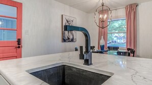 Une cuisine avec un plan de travail en marbre, un évier noir, une porte rouge, un robinet bleu, un lustre et une fenêtre ornée de rideaux roses.