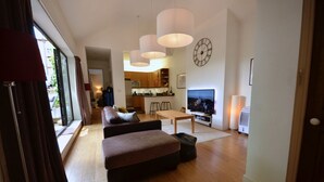 A modern living room with a sofa, ottoman, and a TV. There is a kitchen in the background with wooden cabinets and a microwave. - Spacious Open Plan House in Central Windsor. Dog Friendly + Off Street Parking! (Windsor)