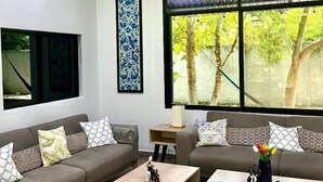 Een moderne woonkamer met een grijze bank, twee houten salontafels en een houten stoel. Op de bank liggen kussens met een patroon en aan de muur hangt een lijst met kunstwerk.