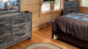 Une chambre avec des murs en bois, une grande fenêtre, un lit, une commode et un téléviseur.