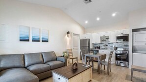 Una sala de estar moderna con un sofá gris, una mesa de centro de madera y una zona de comedor con mesa y sillas. La cocina cuenta con armarios blancos, un refrigerador de acero inoxidable y un horno empotrado.