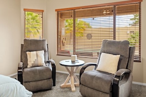 Two grey reclining chairs with cushions, a small round table with a decorative item, and a window with blinds.