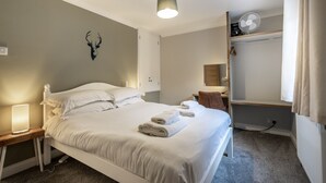 Un lit bien fait, avec des serviettes pliées, une lampe de chevet, une décoration murale représentant une tête de cerf, un ventilateur de plafond et une étagère intégrée avec un miroir et des rangements.
