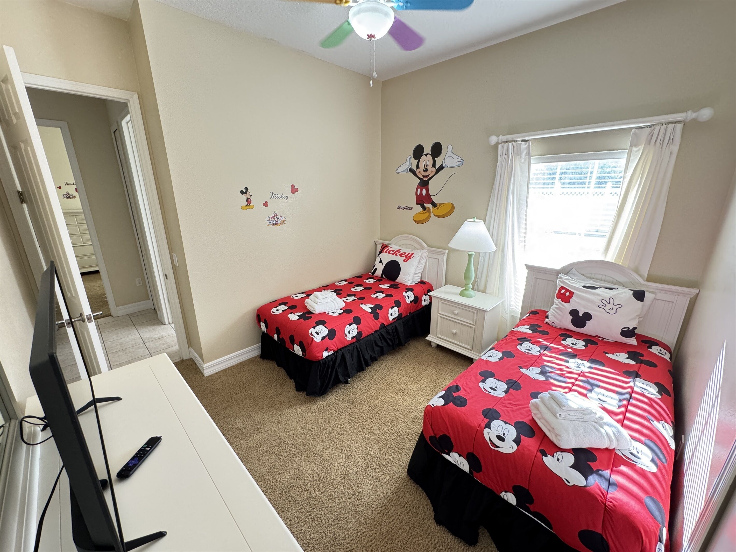 A bedroom with two beds, each with Mickey Mouse-themed bedding, a ceiling fan, a nightstand with a lamp, and a window with curtains.