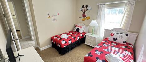 A bedroom with two beds, each with Mickey Mouse-themed bedding, a ceiling fan, a nightstand with a lamp, and a window with curtains.