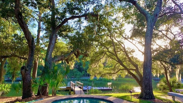 A pool area with a wooden deck, surrounded by trees and a body of water. - Exquisite Home w/ Private Pool & Dock Hilton Head (Hilton Head Island)