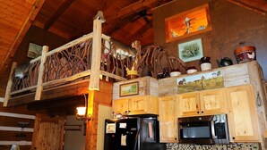 Une cuisine rustique avec des meubles en bois, un réfrigérateur noir, un four à micro-ondes et des objets décoratifs, notamment une sculpture en bois et des tableaux encadrés accrochés au mur.