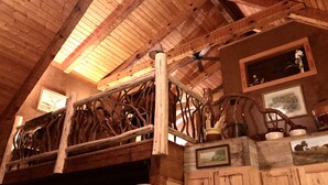 Un intérieur rustique avec des éléments en bois apparents, notamment des poutres, un escalier en bois et des œuvres d’art encadrées qui ornent les murs.