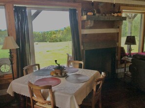 A dining area with a table set for two, a fireplace, and a view of a grassy field through a window. - Hand hewn log cabin with beautiful views of the Blue Ridge Mountains  (Roseland)