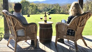Deux chaises en osier sur une terrasse en bois, avec une table placée entre elles, une bouteille et deux verres de vin rouge.