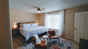 A bedroom with a bed, two bedside lamps, a ceiling fan, a window with curtains, a door, and a patterned rug. - The Happy Cactus Home (Kanab)