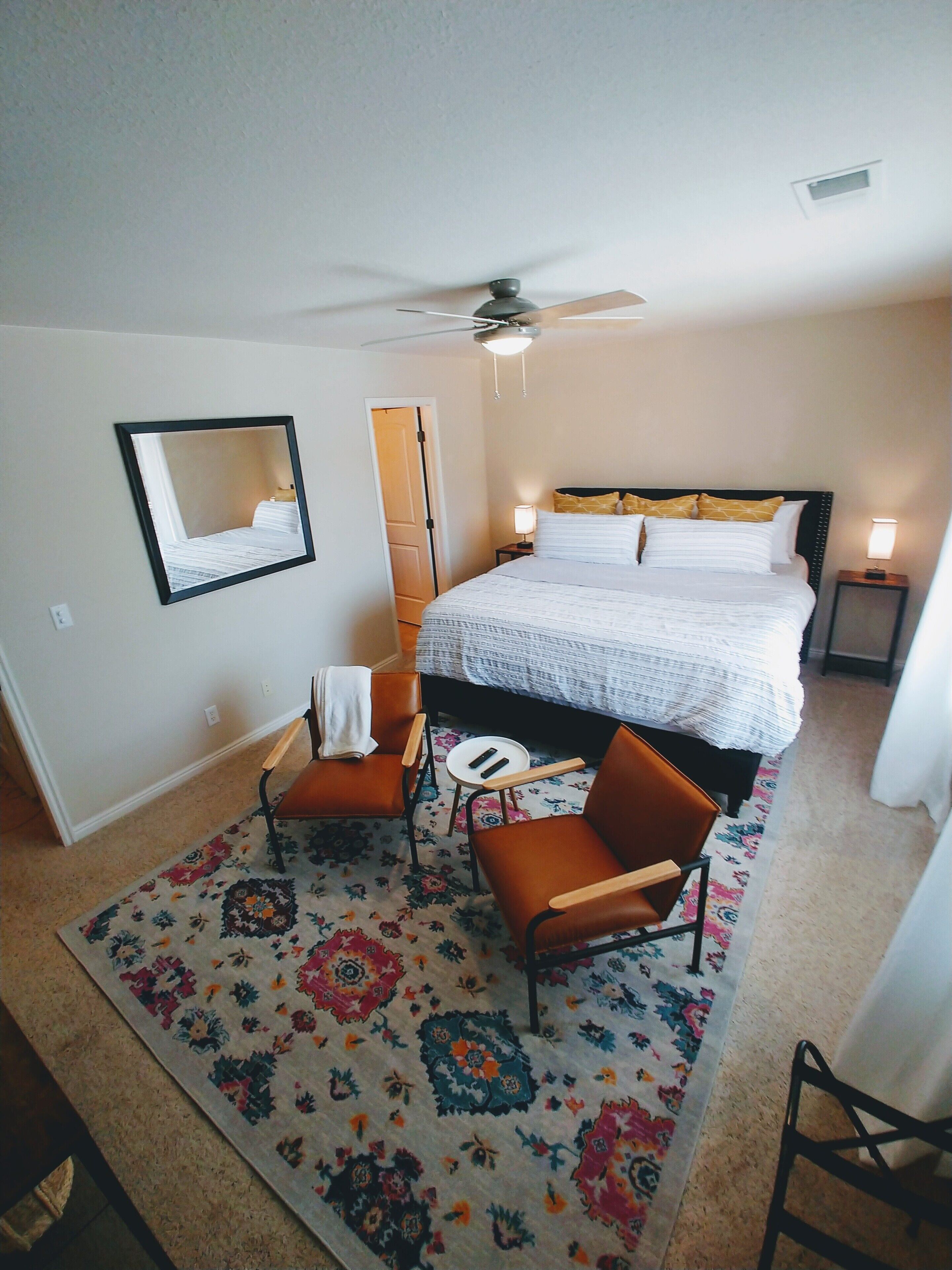 A bedroom with a bed, two chairs, a small table, a ceiling fan, and a mirror on the wall.