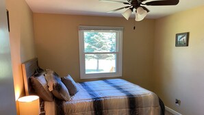 A bedroom with a bed, a nightstand, a lamp, a ceiling fan, a window, and a framed picture on the wall.