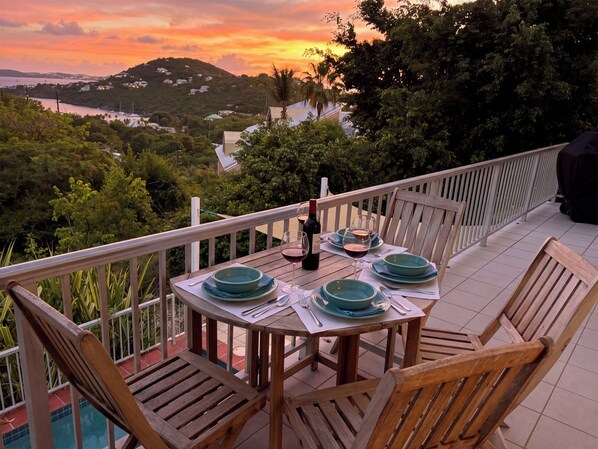 An outdoor dining area with a table set for four, surrounded by wooden chairs, overlooking a scenic view of a coastline and hills.