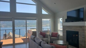 A spacious living room with a large window overlooking a lake, a fireplace, and a flat-screen TV mounted on the wall.