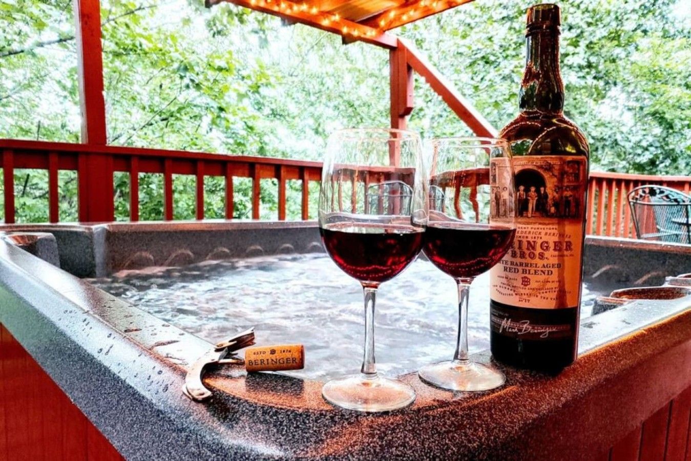 A bottle of wine and two glasses on a stone ledge with a wooden railing and greenery in the background.