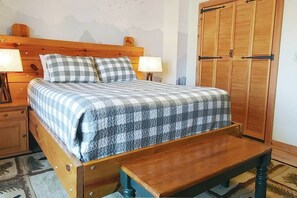 Una cama con una colcha a cuadros, cabecera de madera, lámparas de noche, un banco de madera y un armario con puerta de madera.