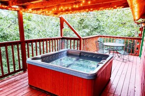 Un bain à remous sur une terrasse en bois, avec une table et des chaises, le tout entouré d’une rambarde et illuminé par des guirlandes lumineuses.
