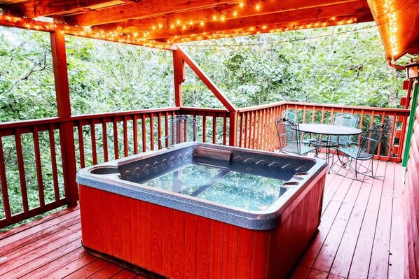 A hot tub on a wooden deck with a table and chairs, surrounded by a railing and string lights.
