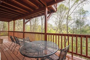 Une terrasse extérieure couverte, dotée d’une table ronde et de chaises, entourée d’une rambarde en bois, et offrant une vue sur une zone boisée.